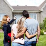 Real,Estate,Agent,Woman,With,Clients,Near,New,House.